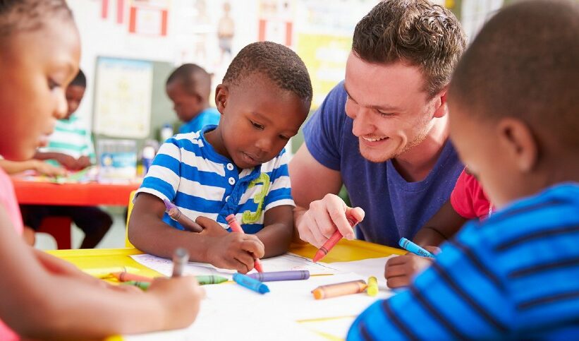 An early childhood education teacher looks on as his young students draw with crayons.