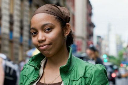 A young women wearing a green jacket stands on a city street and looks directly at the camera with a neutral expression.