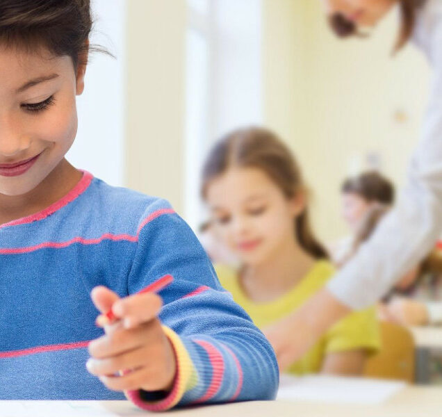 A young elementary school student smiles and holds a writing utensil at their desk while an instructor instructs another student in the background.