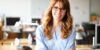 A person with long, auburn hair and glasses smiles for the camera while standing in a school classroom.