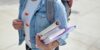 A student wearing a denim jacket carries books, binders and a backpack.