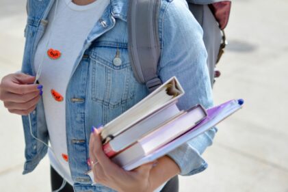A student wearing a denim jacket carries books, binders and a backpack.