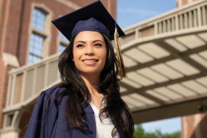 A person wears a graduate cap and gown and half-smile looks into the middle distance contemplating her life choices.